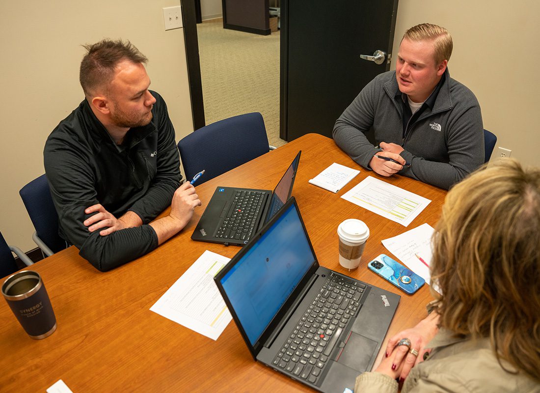 Read Our Reviews - View of Three Synergy Insurance Employees Sitting Around a Wooden Table with Open Laptops in a Small Conference Room During a Business Meeting